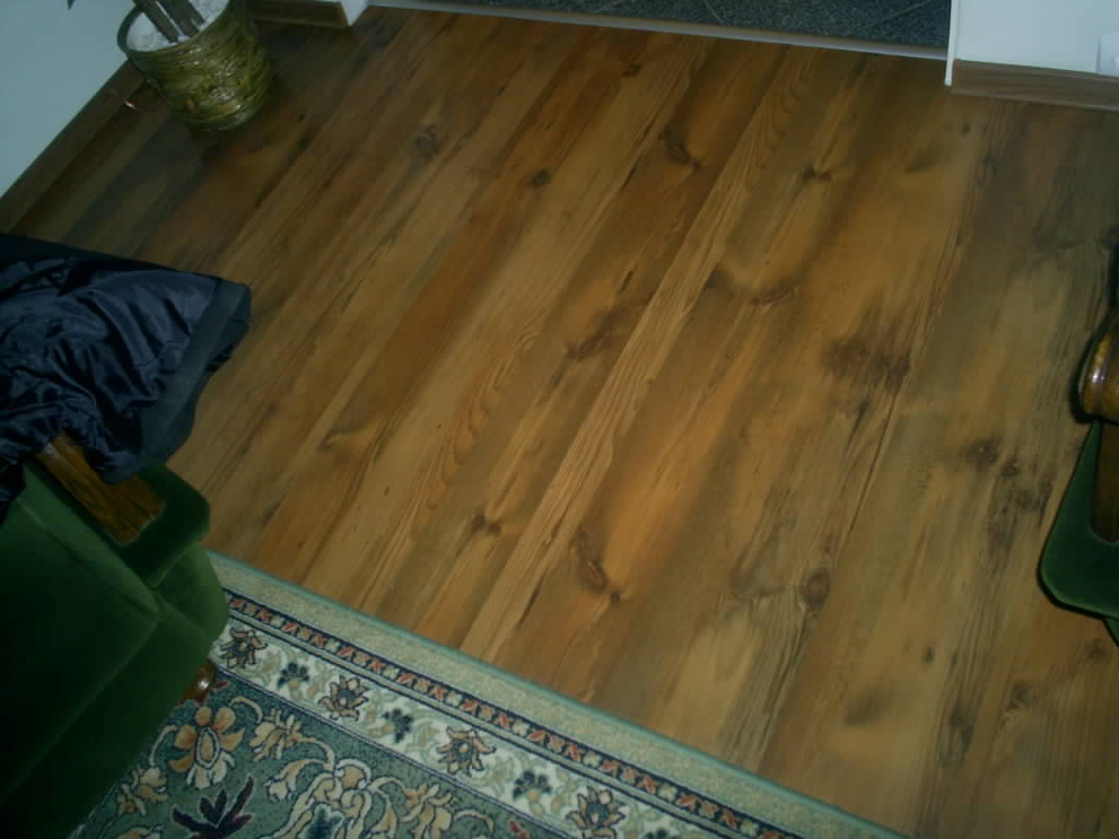 Ułożona pod kątem, lakierowana podłoga drewniana z widocznymi słojami, fragment zielonego dywanu z ornamentami, część zielonego fotela i rośliny doniczkowej w plecionym koszu.